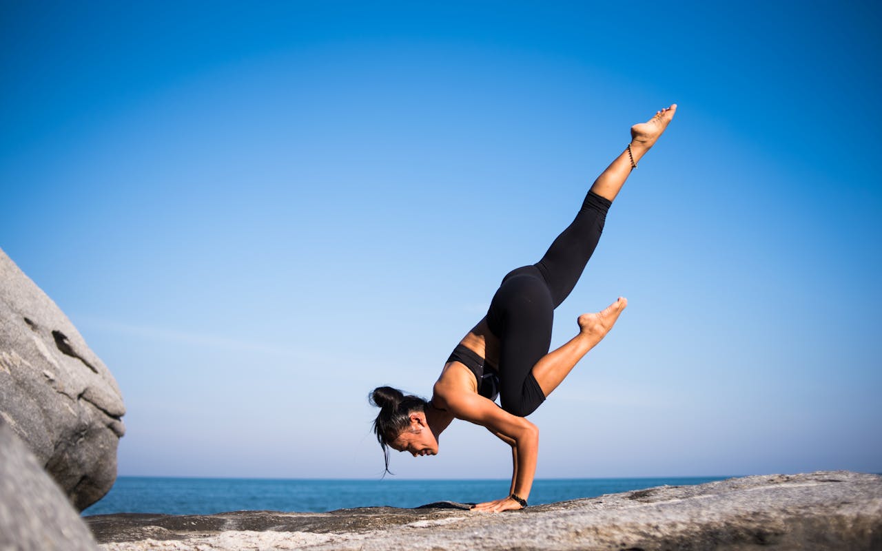 services-02 Woman performing a yoga pose on a rocky beach with ocean view and clear blue sky.